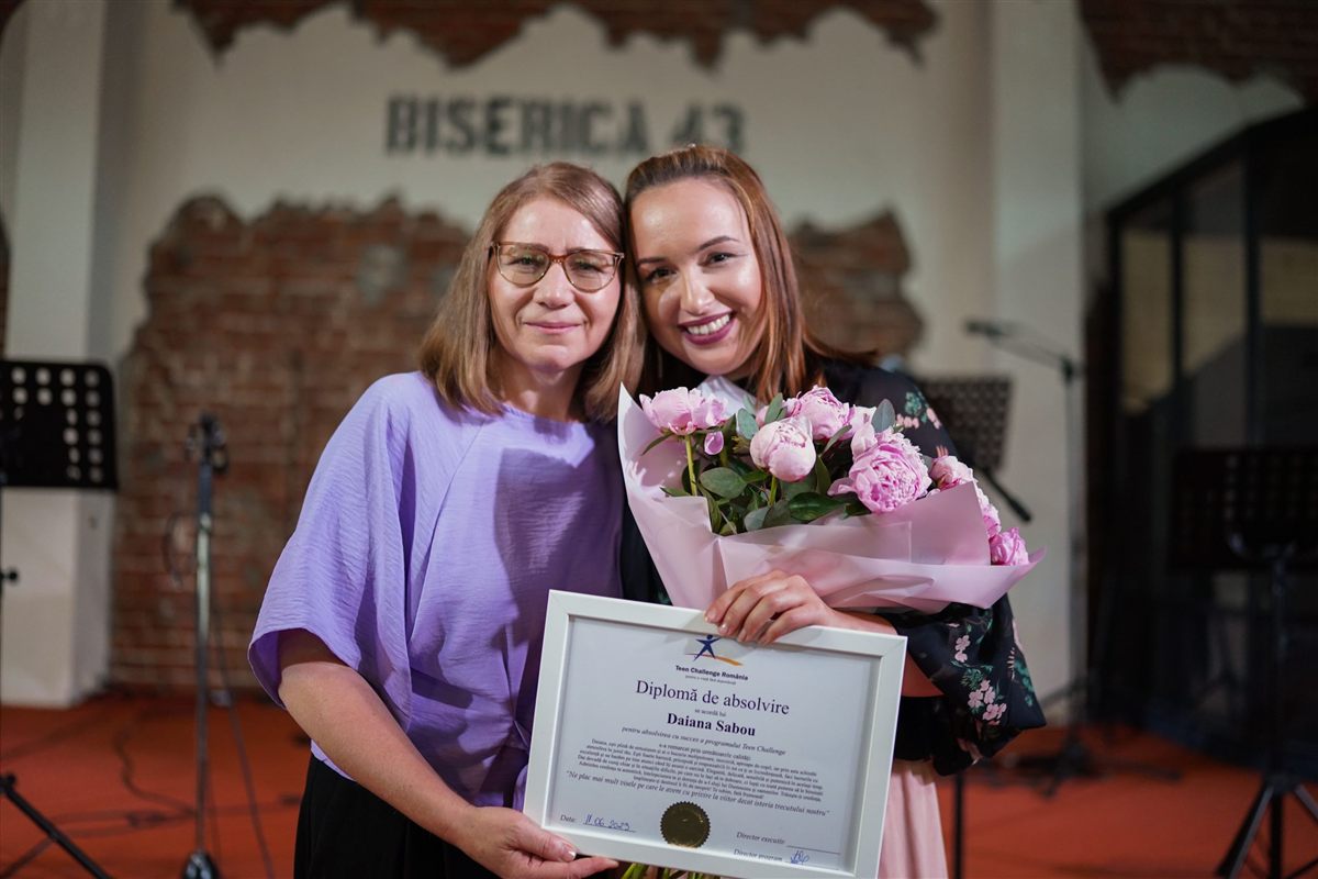 O mamă alături de fiica ei la absolvirea programului, cu diploma și un buchet de flori în mâini